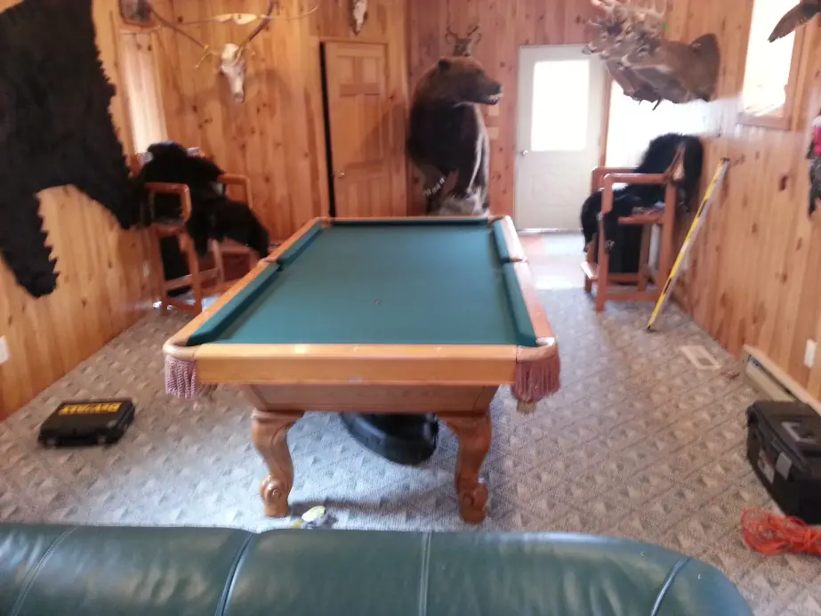 A green felt table with light oak rails and carved legs in a pine-paneled room hung with mounted wildlife