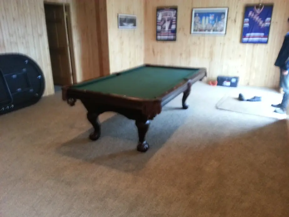A dark-legged pool table with green felt in a pine-paneled rec room