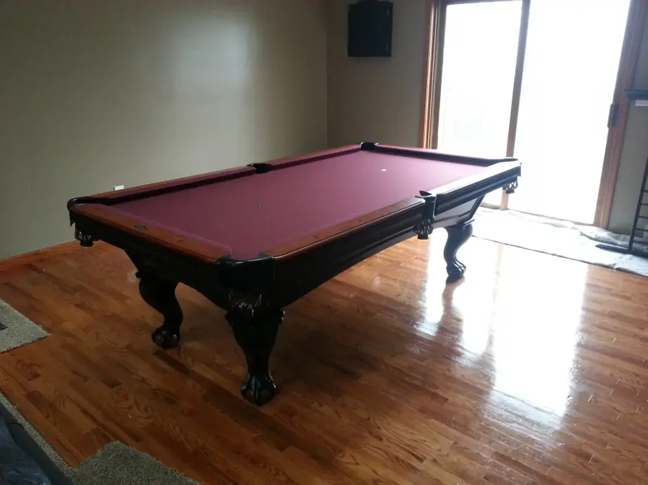 A pool table with wine-colored felt and dark carved legs on a wood floor by a window